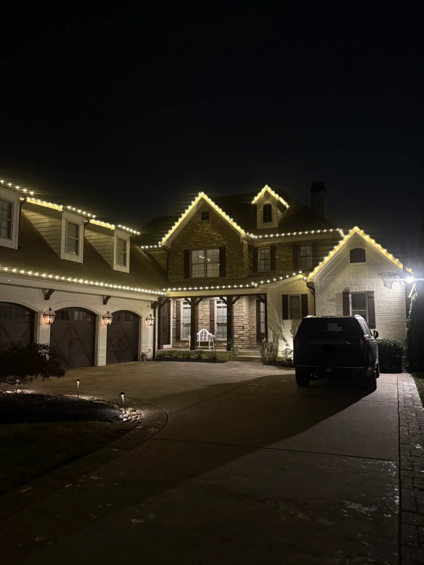 Warm white lights outlining a stone home’s roof peaks, dormers, and porch